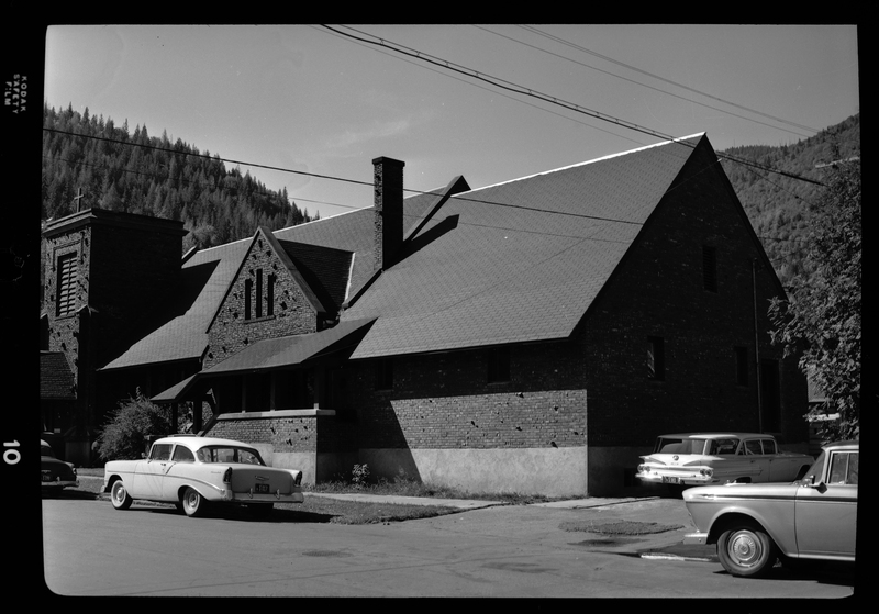 Photo of the exterior of an Episcopal Church in Wallace, Idaho. There are several cars parked along the road and in the alley next to the church.