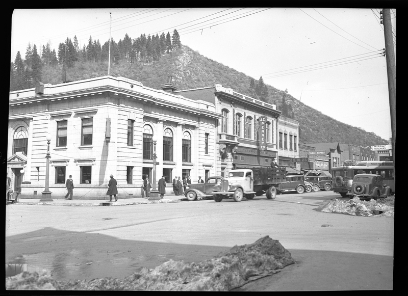 Photo of downtown Wallace, Idaho that apparently appeared in Household Magazine. The J. C. Penney Company store and several other buildings are visible. There is a truck driving down the street that has people walking on it and more cars parked on the sides of the roads. There are also some mostly melted piles of snow next to the roads.