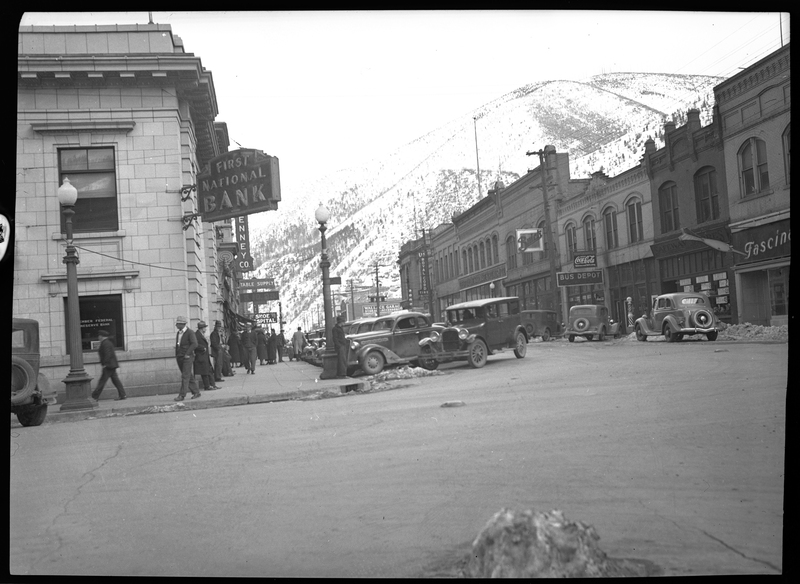 Photo of downtown Wallace, Idaho that apparently appeared in Household Magazine. The First National Bank and the Bus Depot buildings are visible, as well as many other stores. People can be seen walking the sidewalks, and there are cars parked on the sides of the road. There are also some mostly melted piles of snow next to the roads.
