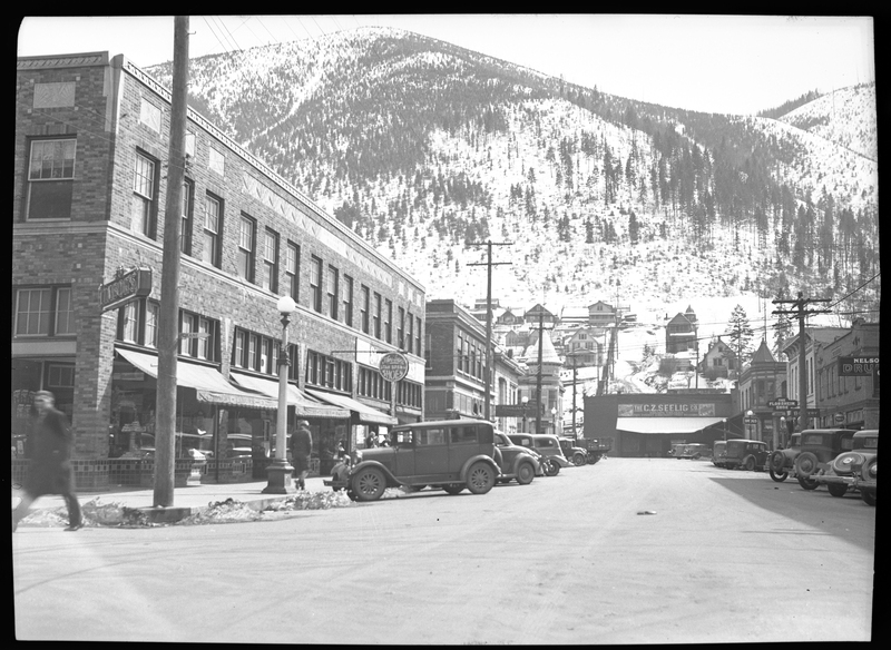 Photo of downtown Wallace, Idaho that apparently appeared in Household Magazine. Tabor's and C. Z. Seelig stores are both visible, as well as others. People walk on the sidewalks and cars are parked on either side of the road. There is snow covering the mountain in the background that is behind the city.