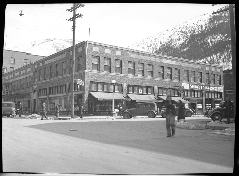 Photo of downtown Wallace, Idaho that apparently appeared in Household Magazine. Stone's Food Stores and Tabor's are visible, as well as a few other shops. People are walking on the sidewalks and across roads, and there are cars parked on the sides of the road in front of the stores. There are also a few piles of melted snow on the ground next to the roads.