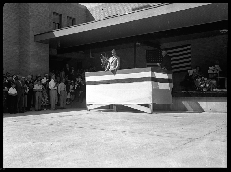 An unidentified man stands on a podium to address a crowd at the dedication of the Wallace Post Office. Part of the crowd is visible standing next to the wall, and another man stands on the stage behind the podium. There is an American flag hung up on the wall behind the man addressing the crowd.