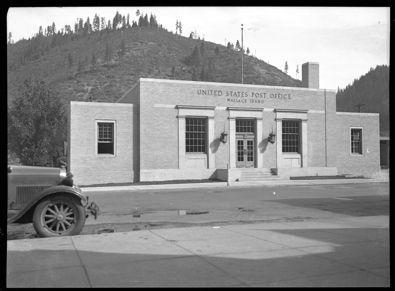 Photo of the exterior of the Wallace Post Office building. The signs on the building read "United States Post Office; Wallace Idaho." The front of car that is parked on the side of the road is just barely visible.