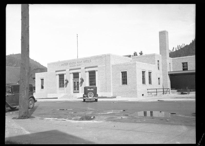 Photo of the exterior of the Wallace Post Office building. The signs on the building read "United States Post Office; Wallace Idaho." Two cars are parked on the side of the road near the post office.