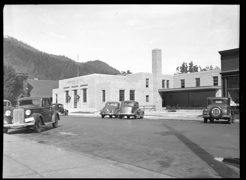 Photo of the exterior of the Wallace Post Office building. The signs on the building read "United States Post Office; Wallace Idaho." Six cars are parked on the side of the road near the post office.