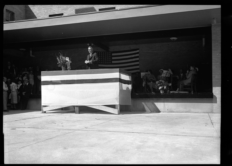 An unidentified man stands on a podium to address a crowd at the dedication of the Wallace Post Office. Part of the crowd is visible standing next to the wall. There is an American flag hung up on the wall behind the man addressing the crowd.