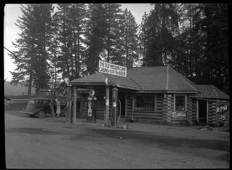 Photo of the Old Mission Camp gas station and convenience store. The sign on top of the building reads, "The Old Mission Camp; Cabins ~ Gilt Top Beer." There are three gas pumps at the front of the building, and a group of people are standing around a car parked outside.