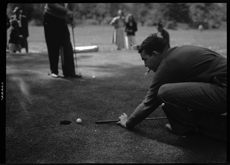 Photo of an unidentified man playing golf at Shoshone Golf Course. The man is knelt on the ground and is preparing to hit the golf ball into the hole. He has a cigarette in his mouth, and other people are standing in the background and watching him.