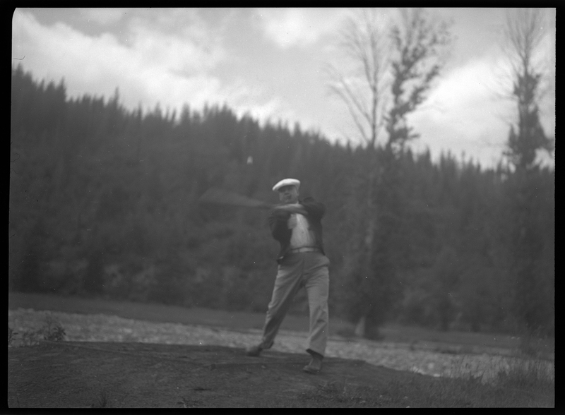 Photo of an unidentified man playing golf at Shoshone Golf Course. The man is mid swing, though he's holding the club like a baseball bat and attempting to hit a flying golf ball.