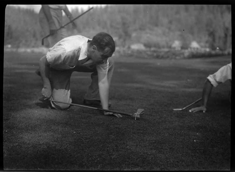 Photo of an unidentified man playing golf at Shoshone Golf Course. The man is knelt on the ground and holding a golf club in his hand, poking it against a small bag on the grass. There is another person knelt on the ground that is mostly cut off from the picture, and another standing in the background.