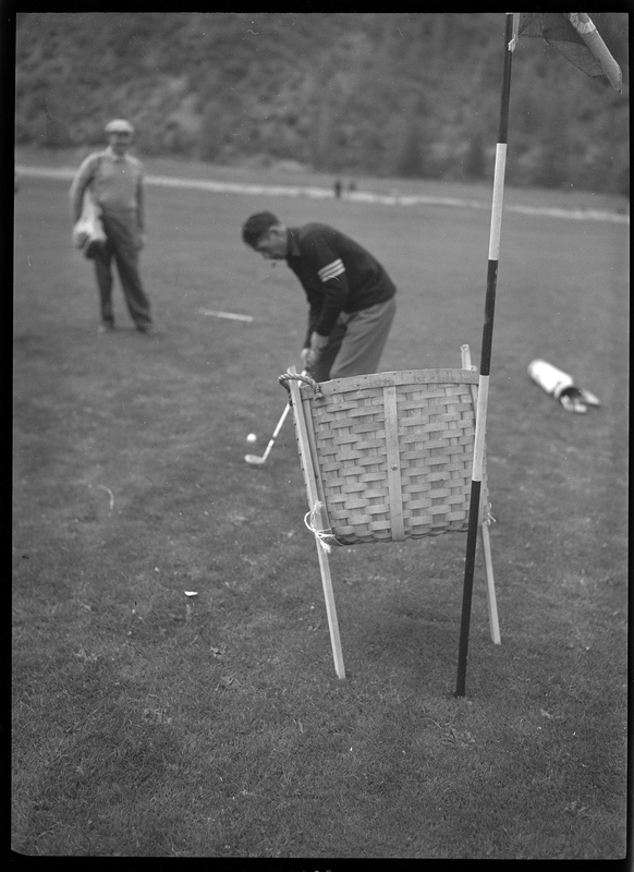 Photo of an unidentified man playing golf at Shoshone Golf Course. The man is preparing to hit the golf ball on the ground in front of him with the club while another man stands off to the side and watches. There is a basket between the man golfing and the photographer.