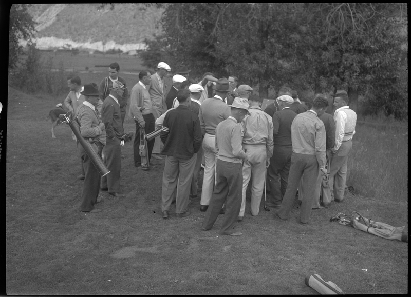 Photo of a group of men huddled together at the Shoshone Golf Course. A couple of the men have golf bags with their clubs in them, but most are just holding a single golf club. None of them are looking at the camera.