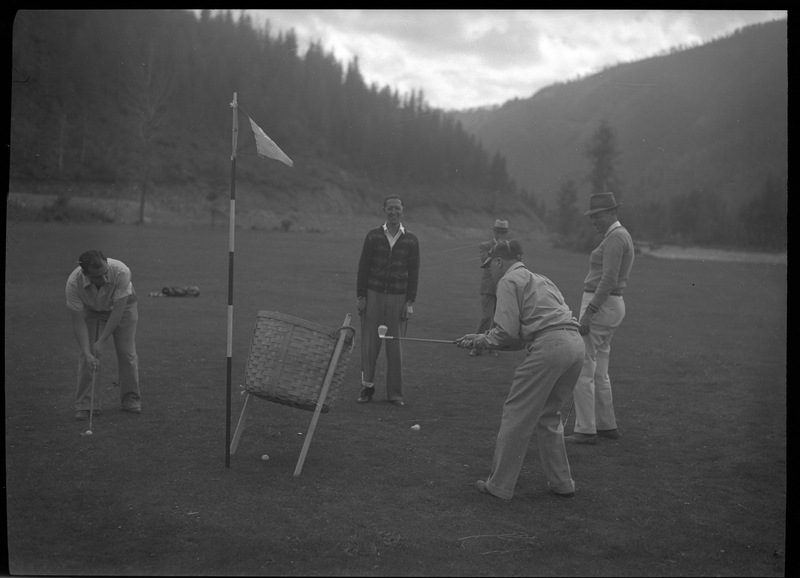 Photo of five men standing near each other while two of them are playing golf. The three not playing are watching and laughing.