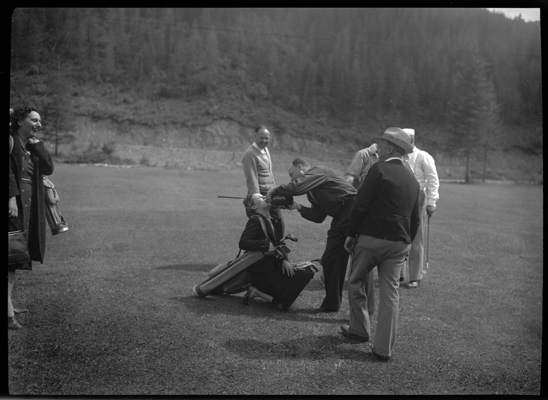 An unidentified man is pouring a drink into a woman's mouth who is knelt on the ground in front of him. She has a bag over her shoulder that is holding golf clubs. The men and women around them are watching and laughing.