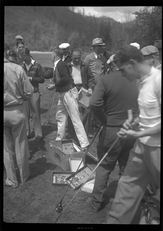 A group of men stand together around a box at the Shoshone Golf Course. Most of the men are holding golf clubs.