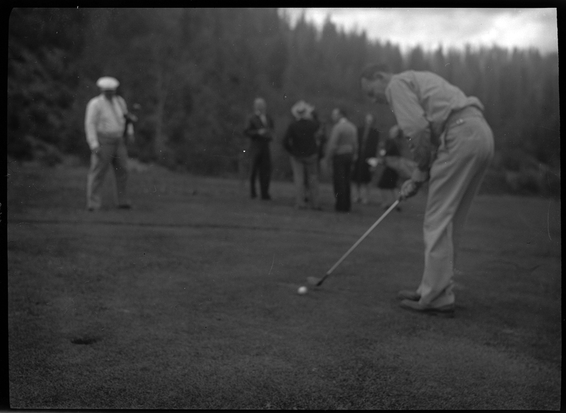 Photo of an unidentified man playing golf at Shoshone Golf Course. The photo is out of focus, but a man can be seen preparing to hit a golf ball for the hole. There are other people standing together in the background.