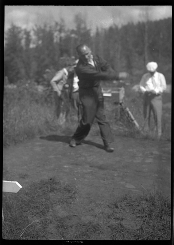 Photo of an unidentified man playing golf at Shoshone Golf Course. The man is mid swing with the golf club and there are a few people standing behind him. The photo is out of focus.