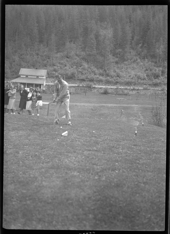 Photo of an unidentified man playing golf at Shoshone Golf Course. He is preparing to hit the golf ball in front of him while a group of women watch him in the background.