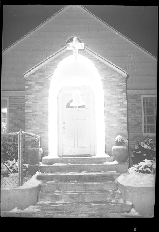 Photo of the front of the Chemadurow house. The light above the front door is bright and leaves the area washed out, but there appears to be a cross above the door. There is snow on the ground and steps leading up to the building.