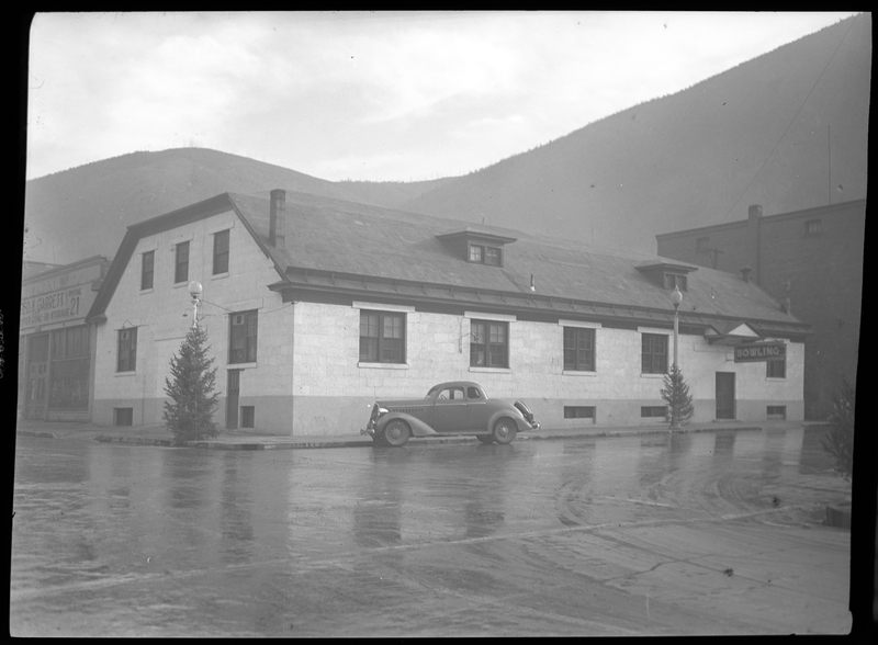 Photo of the exterior of the Wallace Bowling Alley in Wallace, Idaho. There is a car parked on the side of the road next to the building. The street appears to be flooded.