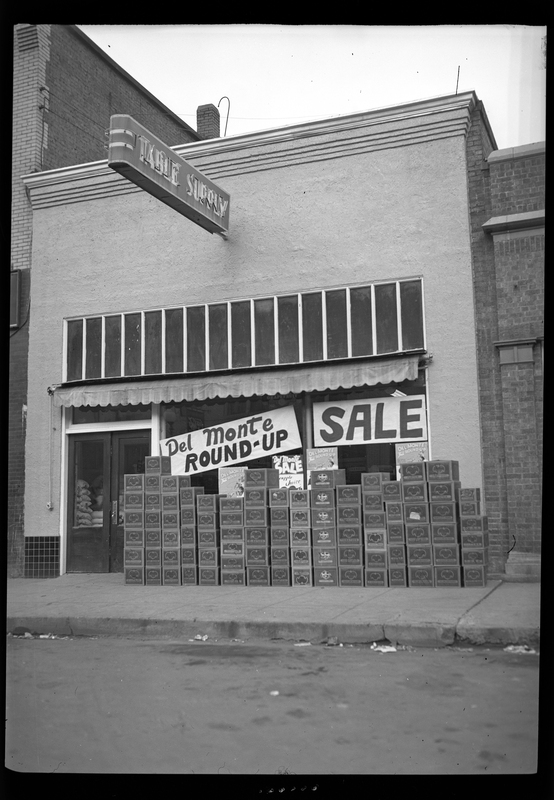 Photo of the Del Monte Round Up sign that is hung in the window of the Table Supply store. There is another sign in the window that reads "sale." On the ground outside of the building there is a line of boxes stacked up.
