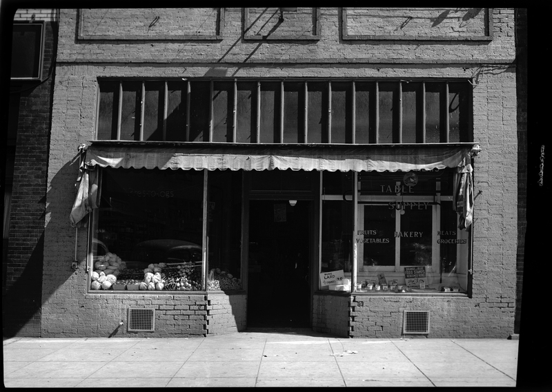 Photo of the storefront of Table Supply. There are two window displays on either side of the front door and one of the windows has several decals that read, "Table Supply," "Fruits," "Bakery," Meats," "Vegetables," "Groceries." 