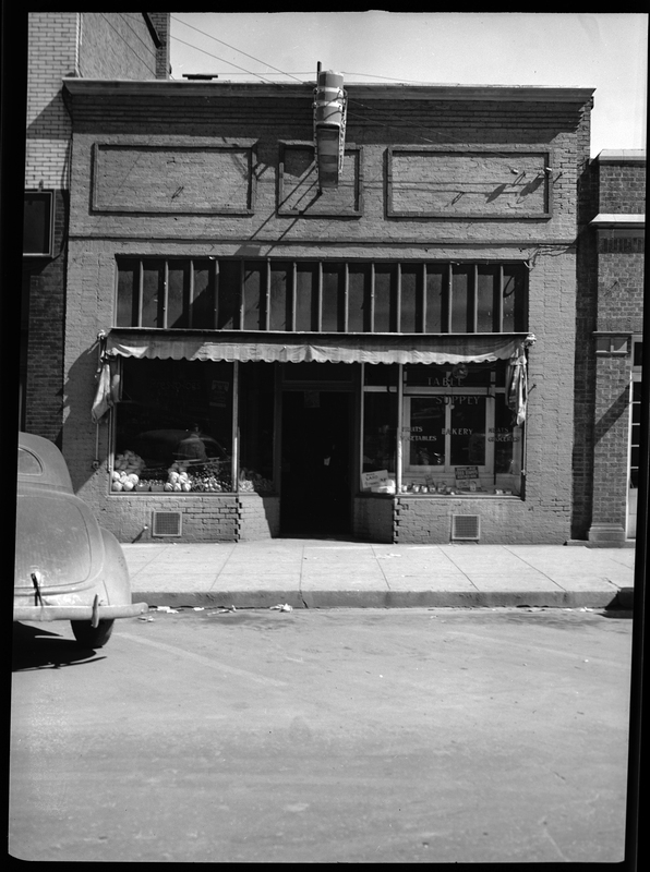 Photo of the storefront of Table Supply. There are two window displays on either side of the front door and one of the windows has several decals that read, "Table Supply," "Fruits," "Bakery," Meats," "Vegetables," "Groceries." There is a car parked in front of the building.