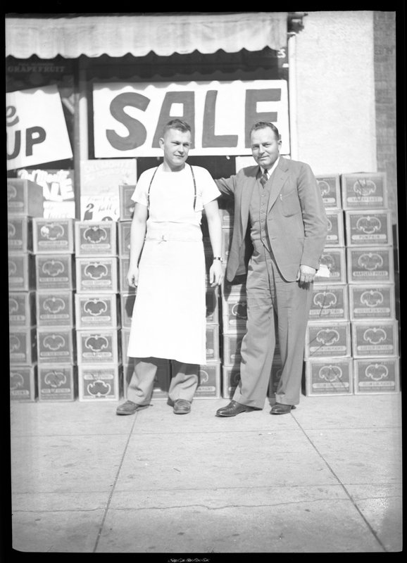Two unidentified men stand in front of several stacks of boxes with a sign behind them that reads "sale." One man is wearing a suit and the other appears to be wearing an apron.