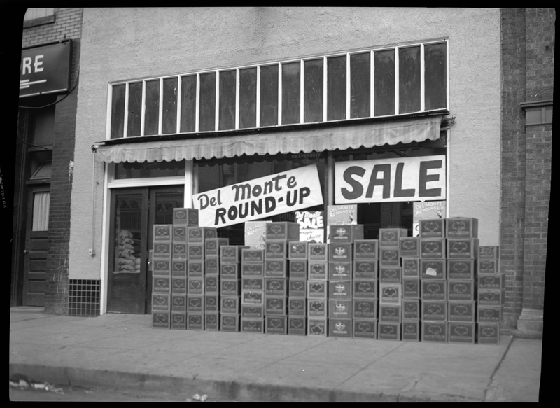 Photo of the Del Monte Round Up sign that is hung in the window of the Table Supply store. There is another sign in the window that reads "sale." On the ground outside of the building there is a line of boxes stacked up.