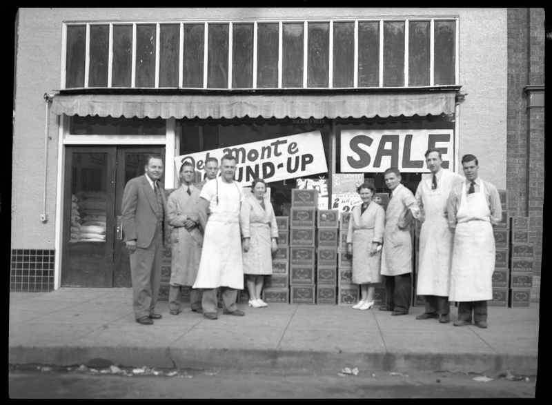 Photo of nine unidentified men and women standing in front of a storefront that has two signs hanging in the windows, one of which reads, "Del Monte Round-Up," and the other reads, "Sale." They are all stood in front of a line of stacked boxes and most of them are wearing aprons.