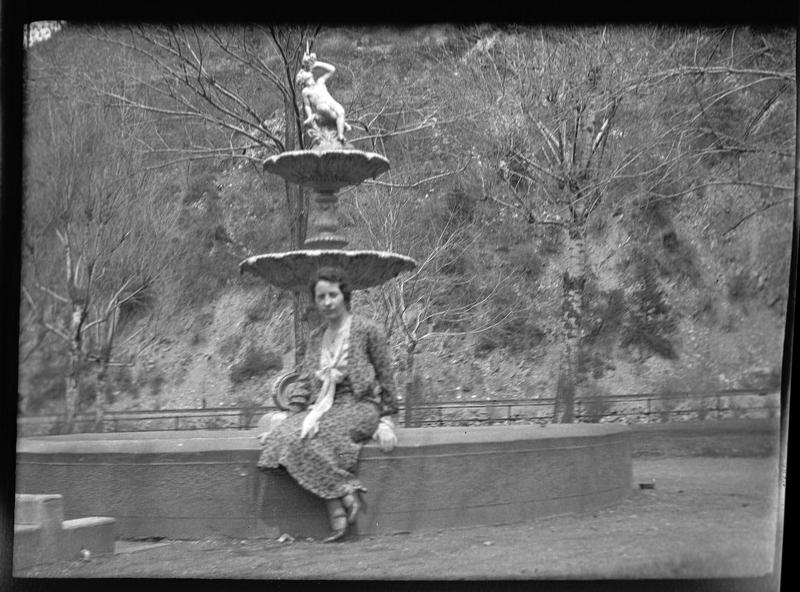 Photo of an unidentified woman who is sitting on the ledge of a fountain in the Wallace, Idaho city park. She is wearing a dress and is looking directly at the camera. 