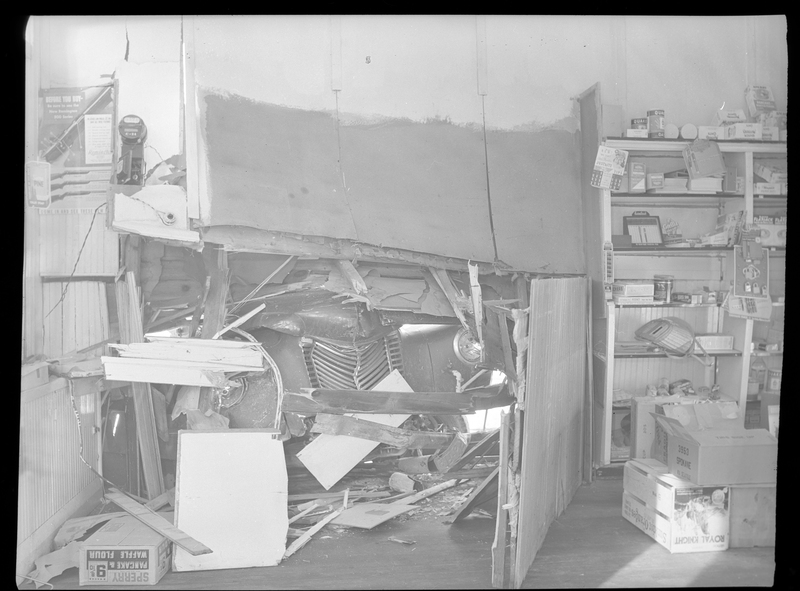 Photo of a truck that was crashed into the side of the General Food Store, taken from the interior of the building. The front and hood of the truck are visible through the hole in the wall, and debris from the impact is all around the room. To the right of where the truck made contact, several shelves with products for sale are visible.