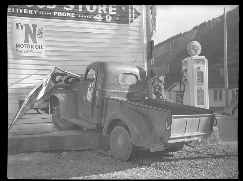 Photo of a truck that was crashed into the side of the General Food Store, damaging the wall and a sign on the exterior. There is a gasoline pump directly to the right of the truck, and a conoco sign fell on the hood. There is a man standing around the corner of the building who is looking at the camera. The license plate on the truck reads "4R652."