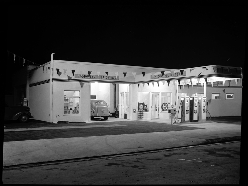 Photo of the Union Oil Dealer building at night. The store is still open with the lights on, and there is a car parked in the garage. Above the garage is a sign that reads, "Stop-Wear Lubrication" and outside there are three gas pumps.
