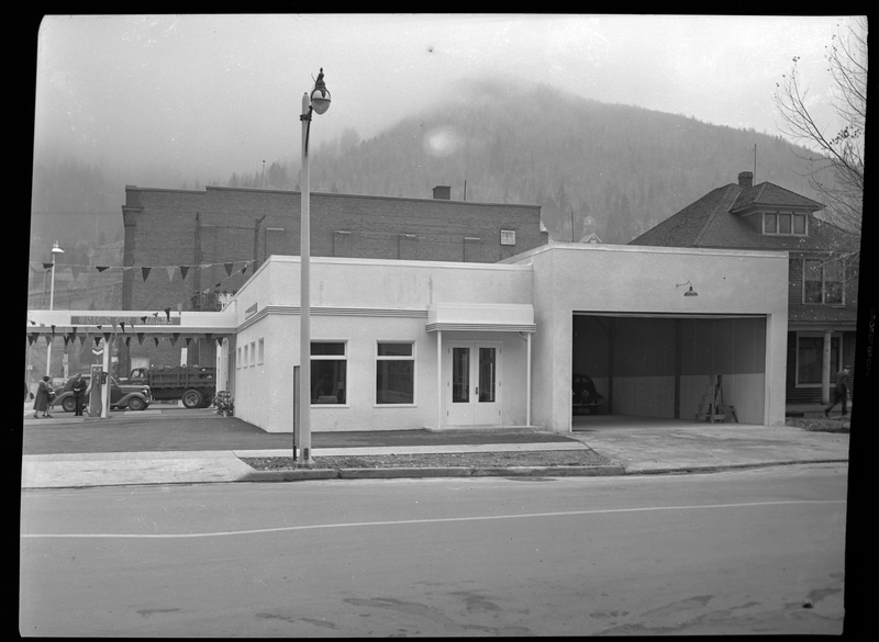 Photo of the Union Oil Dealer building from the side, showing an open garage door that can fit multiple cars. There is one car parked inside it. A few people can be seen on the other side of the building standing near the gas pumps.