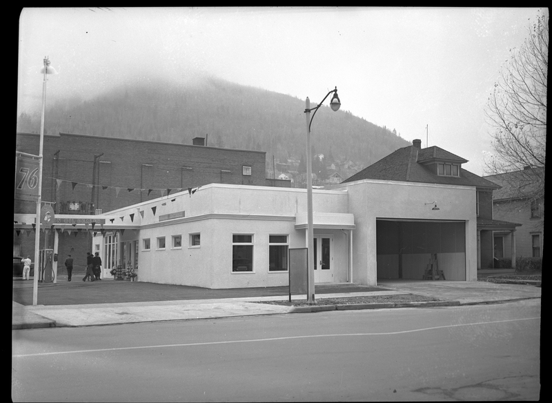Photo of the Union Oil Dealer building from the side, showing an open garage door that can fit multiple cars. A few people can be seen on the other side of the building standing near the gas pumps.