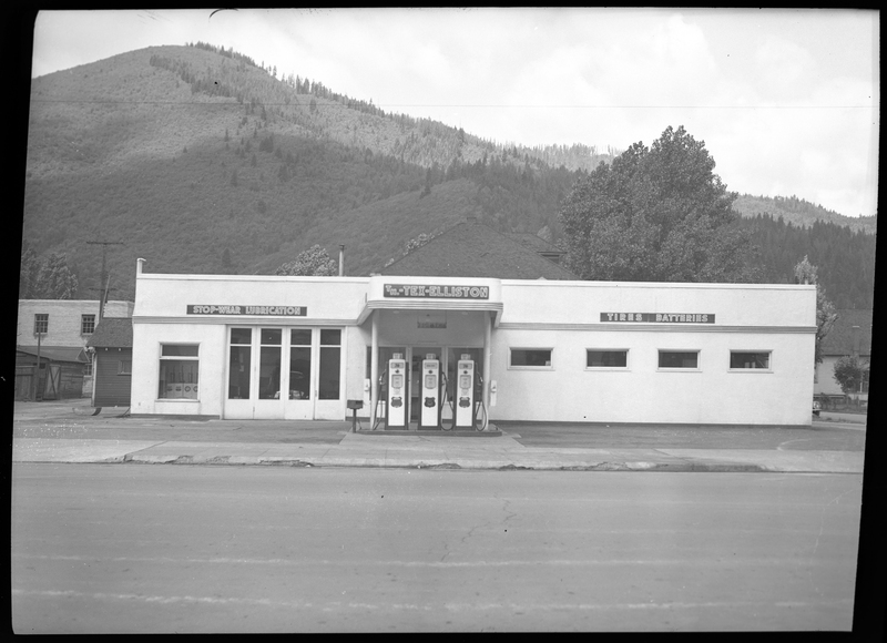 Front of the Union Oil Dealer building. Three gas pumps are in front of the middle of the building with a sign above it that reads "TM.-Tex-Elliston." To the left, there is a garage door that is closed with a sign above it that reads, "Stop-wear lubrication," and to the right there is another sign that reads, "Tires; Batteries."