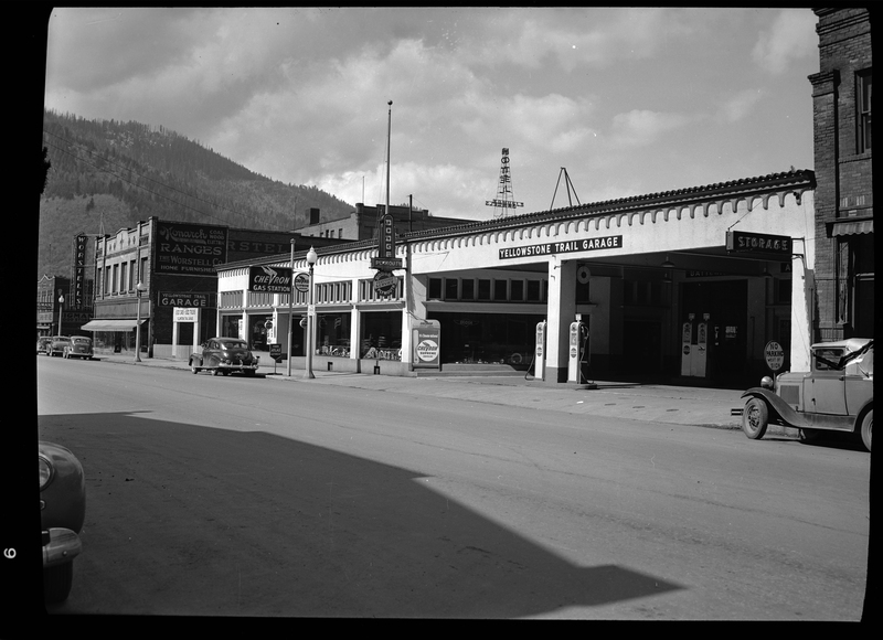 Photo of the Yellowstone Trail Garage (which is now the Shoshone County Jail) in Wallace, Idaho. Several gas pumps are visible under the canopy of the garage, and the Worstells building is visible in the background. Several cars can be seen parked on the side of the road throughout the street.