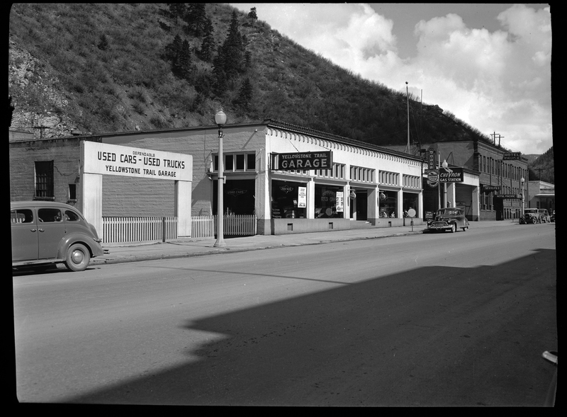 Photo of the Yellowstone Trail Garage (which is now the Shoshone County Jail) in Wallace, Idaho. To the left of the garage building is a sign that reads "Dependable Used Cars - Used Trucks Yellowstone Trail Garage." Cars are seen parked on the side of the road in front of the Yellowstone Trail Garage and further down the street, where the Hotel Pacific is in view.