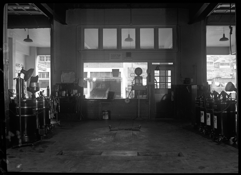 Photo of what is possibly the interior of a gas station, or perhaps the Yellowstone Trail Garage. Two garage doors are visible on the wall the photographer is facing, both of which are open. There are multiple canisters for car repair, some of which read "oil drain" or "lubricant pump." All other labels are not visible and/or illegible.