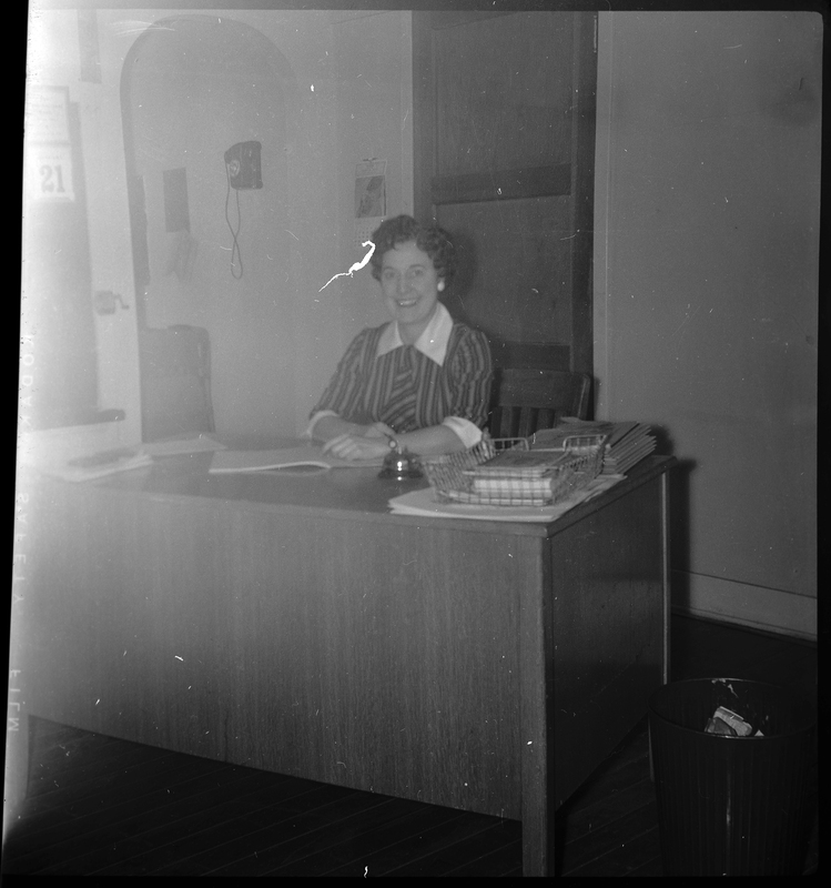 Photo of a woman, probably a teacher, sitting behind a desk at the Opportunity School. She is smiling at the camera and appears to be writing something in a notebook on top of the desk. The negative is slightly damaged, but it does not impede the photo.