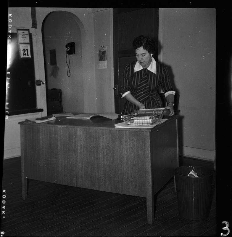 Photo of a woman, probably a teacher, standing behind a desk and adjusting things on top of it. 