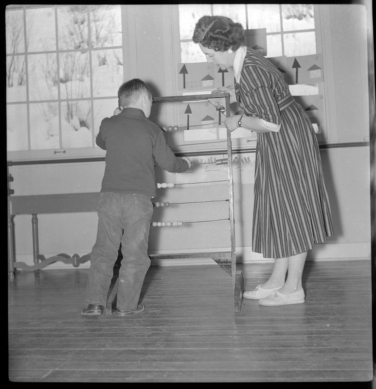 Photo of a woman and a child playing with a device that has beads on wires in the Opportunity School. Neither of them are looking at the camera. The woman is probably a teacher.