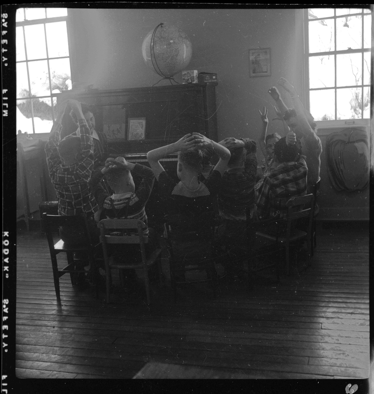 A group of children and two women sit in a circle around a piano in the Opportunity School. Most of the people have their hands raised above their heads and are looking to the woman who is likely the teacher sitting at the piano.