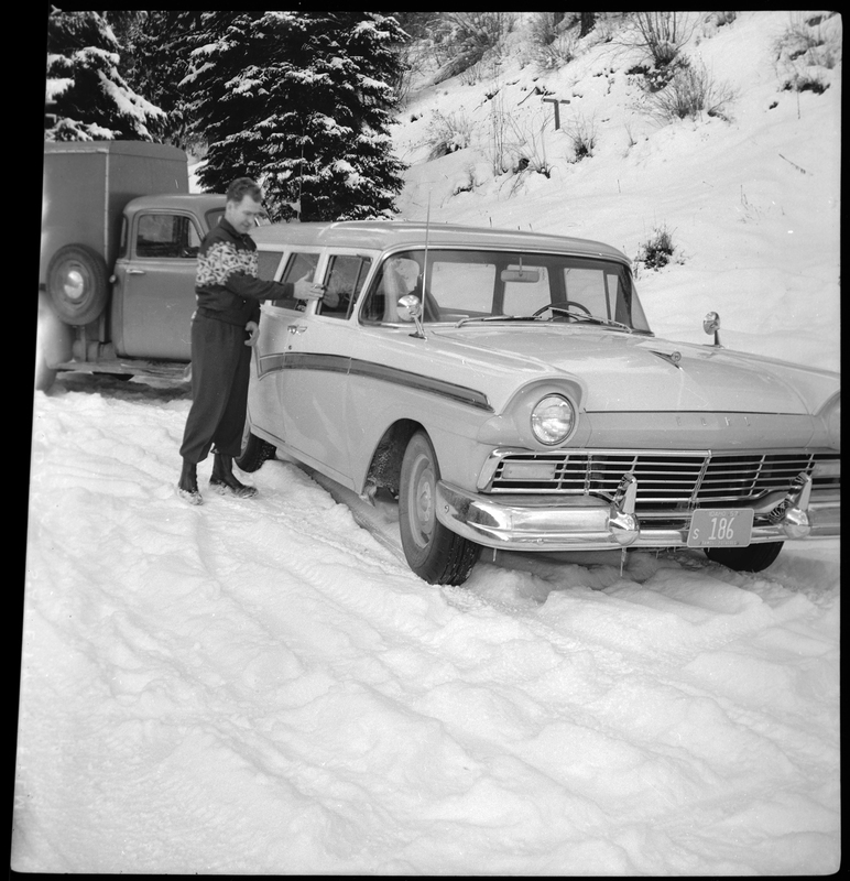 An unidentified man stands next to a car in the snow with a truck in the background. Described as "Opportunity School." The license plate on the car reads, "186."