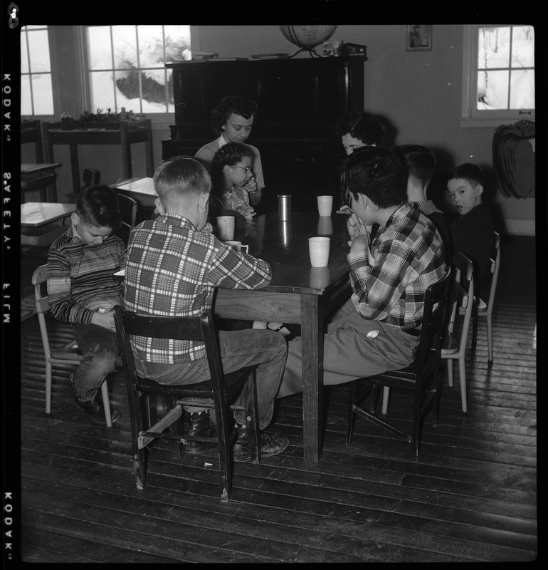 Photo of two women and six children sitting around a table in the Opportunity School. They all have cups in front of them on the table.