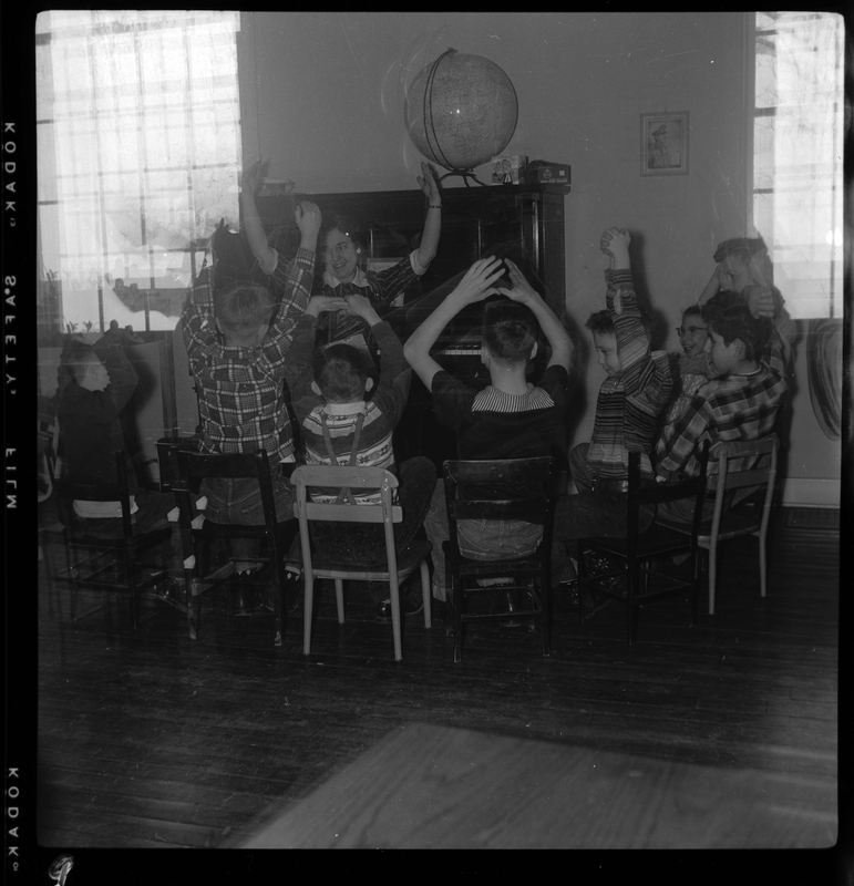A group of children and two women sit in a circle around a piano in the Opportunity School. Most of the people have their hands raised above their heads and are looking to the woman who is likely the teacher sitting at the piano.