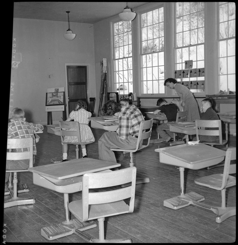 Photo of a classroom within the Opportunity School where several children are sat at desks and working. There is a woman, likely the teacher, helping one of the children.