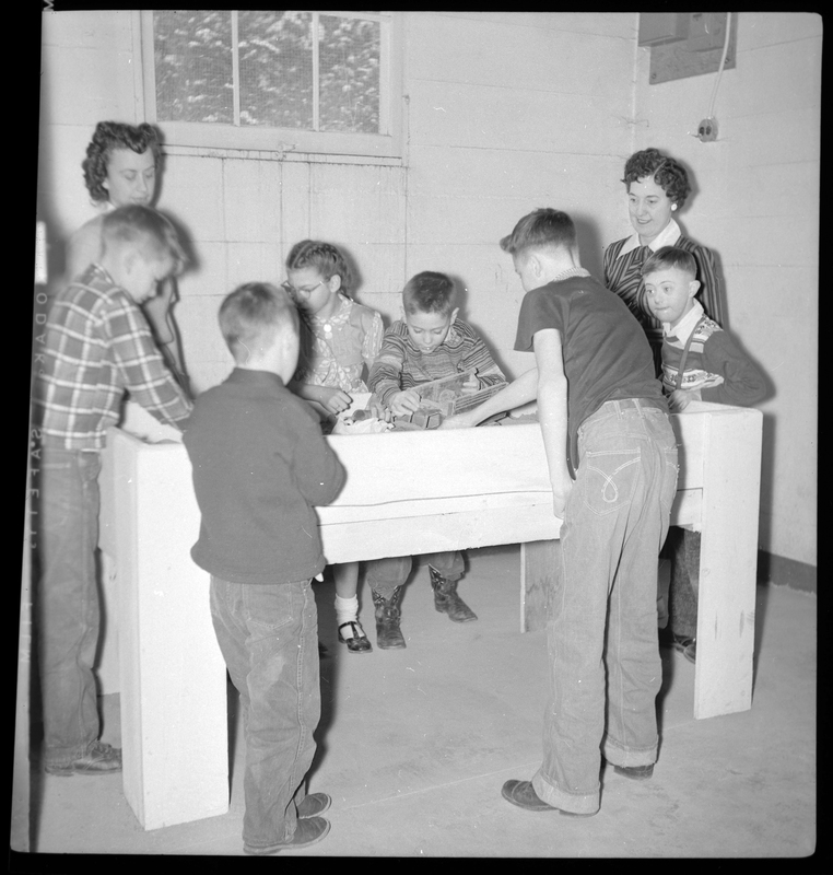 Photo of a group of children and two women, likely teachers, standing around what appears to be a sandbox. The sandbox comes up to the adult's hips and is located inside the building. The children are playing inside of it while the teachers watch.