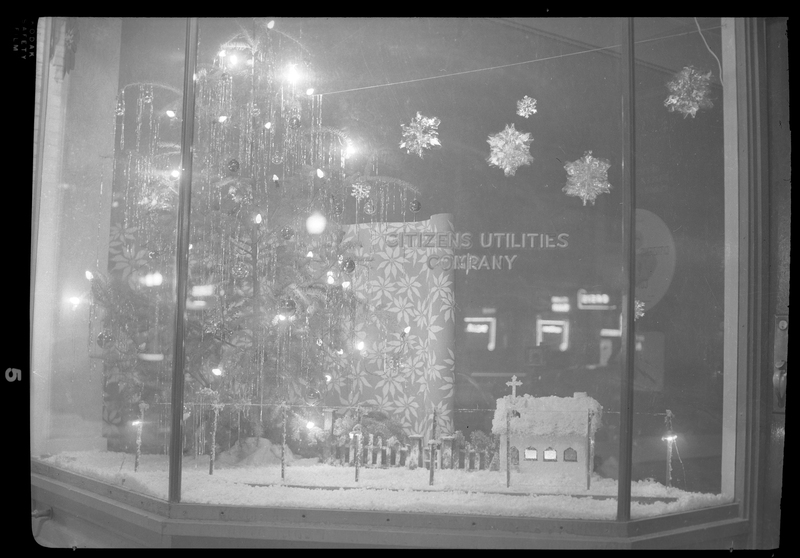 Photo of the Christmas themed window display inside the Citizens Utility building. There is a sparsely decorated Christmas tree and fake snow.